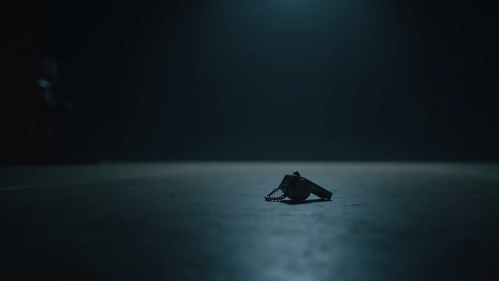 A single small metal whistle lying alone on a wide bare dark floor, small and far from the camera.