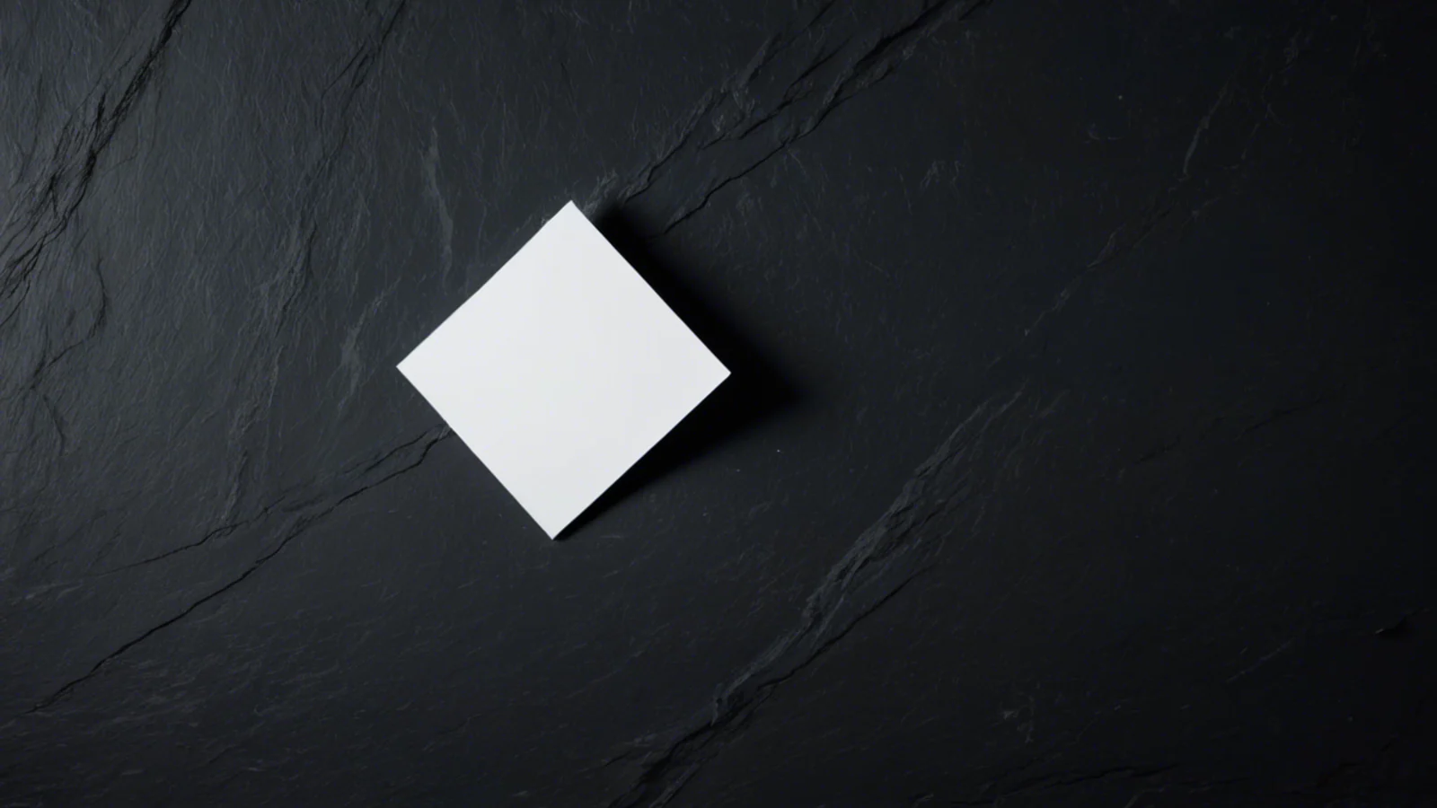 A single small blank white card lying alone on a wide dark slate surface under hard raking light, throwing a long shadow, with deep empty space all around it.